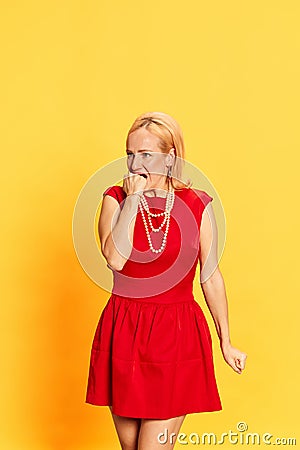 Portrait of scared blond woman in retro clothes biting fist with frightening face on yellow background. Fear, problem Stock Photo