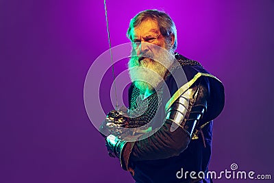 Portrait of senior greybearded man, brave and brutal medieval warrior or knight in armor with sword isolated over purple Stock Photo