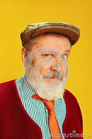 Portrait of senior man in classical clothes, glasses and cap posing over vivid yellow background. Curious, serious look Stock Photo