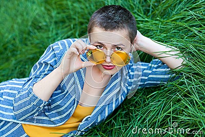Portrait of serious cute young woman with short hair in casual blue striped suit, yellow shirt and eyeglasses lying down on green Stock Photo