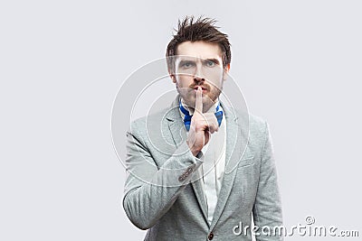 Portrait of serious handsome bearded man in casual grey suit and blue bow tie standing, looking at camera with silence sign Stock Photo