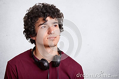 Portrait of serious pensive curly male teenager with curly hair, uses modern headphones for listening favourite music, looks pensi Stock Photo