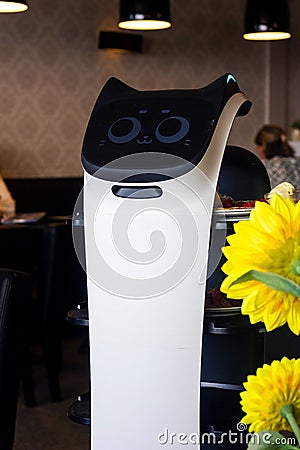 A portrait of a serving robot on wheels with a screen displaying a cute cat face, riding through a restaurant with plates of food Editorial Stock Photo