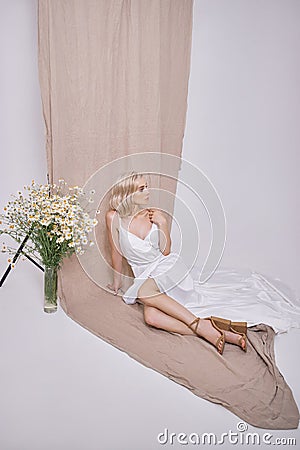Portrait of a sexy blonde in a beautiful white dress near a vase of wild flowers. Romantic girl with beautiful natural makeup Stock Photo