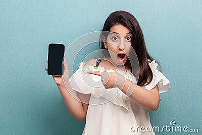 Portrait of shocked beautiful teenager girl in white dress standing, holding phone and pointing finger at display with opened Stock Photo