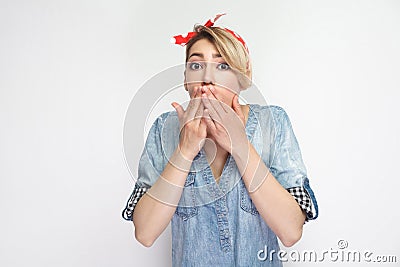 Portrait of shocked beautiful young woman in casual blue denim shirt, makeup and red headband standing and covering her mouth and Stock Photo