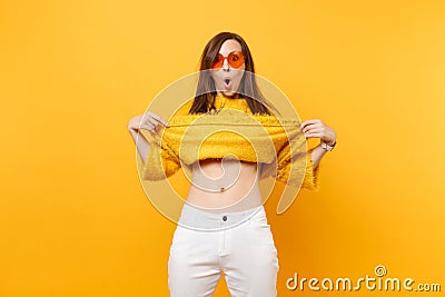 Portrait of shocked funny young woman in heart orange glasses taking off fur sweater, showing belly isolated on bright Stock Photo
