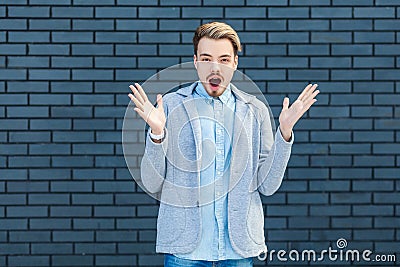 Portrait of shocked handsome young blonde man in casual style standing with raised arms and looking at camera with unbelievable Stock Photo