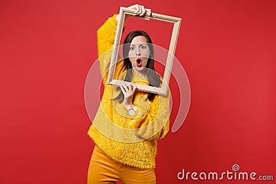 Portrait of shocked young woman in yellow fur sweater keeping mouth wide open, hold picture frame isolated on red Stock Photo