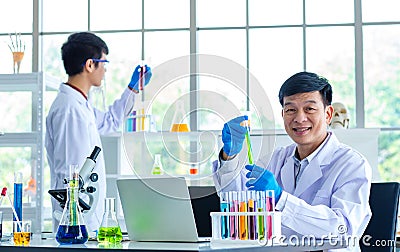 Portrait shot of Asian professional mature male scientist in white lab coat and rubber gloves sitting look at camera smiling Stock Photo