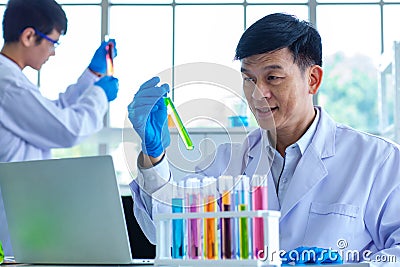 Portrait shot of Asian professional mature male scientist in white lab coat and rubber gloves sitting look at camera smiling Stock Photo