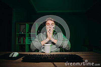 Portrait of a sleepy gamer guy in a headset on his head sitting at a computer with a cup of drink and staring wearily at the Stock Photo