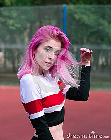 Portrait slim sensual girl cheerleader with pink hair and blue eyes in sportswear posing outdoors near fence at stadium Stock Photo