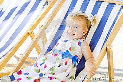 Portrait of smiling adorable blondy baby girl in dress or toddler child relaxing on a sunbed or a deckchair in the city park recre Stock Photo