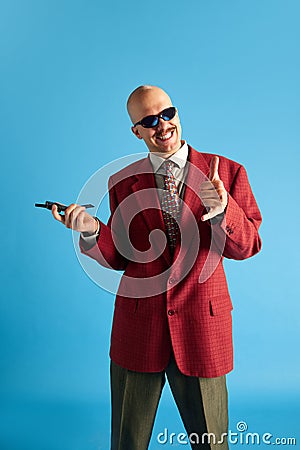 Portrait of smiling balded man with mustache dressed in red retro jacket with vintage mobile in hand over blue studio Stock Photo