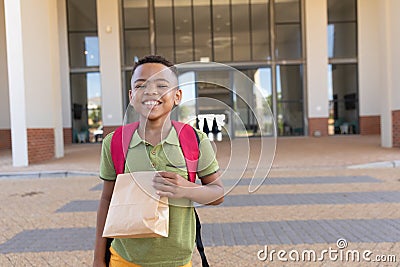 Portrait of smiling biracial elementary schoolboy with packed lunch standing in school campus Stock Photo