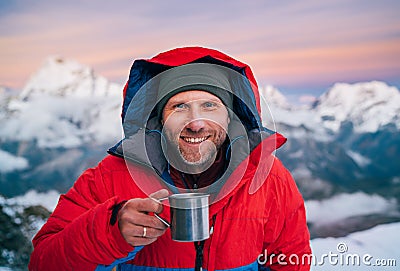 Portrait of smiling at camera high altitude mountaineer dressed red warm dawn jacket holding metal mug of hot tea in with Stock Photo