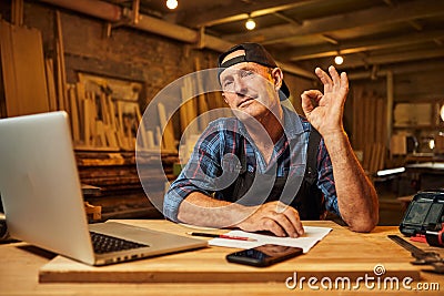 Portrait of smiling carpenter works on the computer and shows sign okay in the workshop Stock Photo