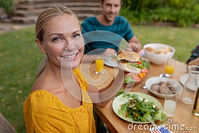 Portrait of smiling caucasian woman holding hamburger eating meal with family in garden Stock Photo