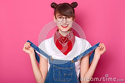 Portrait of smiling female with funny bunches, wears t shirt, denim overalls and bandana on neck, has toothy smile, pulls Stock Photo