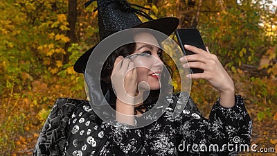 Portrait of a smiling girl in a witch hat and cloak. A girl in a carnival costume is drawn a face for Halloween in the open air. Stock Photo
