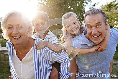 Portrait Of Smiling Grandparents Giving Grandchildren Piggyback Ride Outdoors In Summer Park Stock Photo