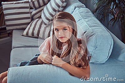 Portrait of a smiling little girl with long brown hair and piercing glance, lying on a sofa at home Stock Photo