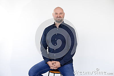 Portrait of smiling middle-aged man wear blue shirt, jeans sitting on stool, interlacing fingers on white background. Stock Photo