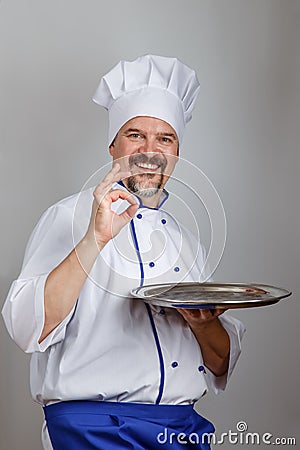 Portrait of a smiling professional chef cook man with a metallic round tray dressed a chef uniform gesturing ok, grey background Stock Photo