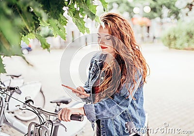 Portrait of smiling red curled long hair caucasian teen girl unlocking bike at Bicycle sharing point using the modern smartphone. Stock Photo