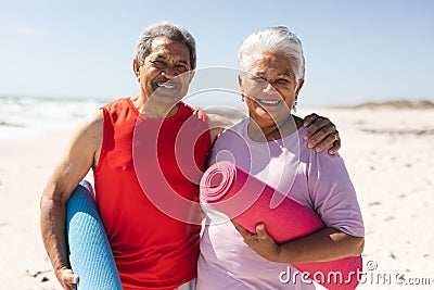 Portrait of smiling senior biracial man with arm around woman holding yoga mats at beach Stock Photo