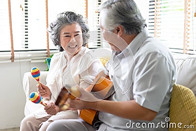 Portrait of smiling senior couple playing guitar singing and her wife holding maracas dancing and sitting on sofa at home. Stock Photo