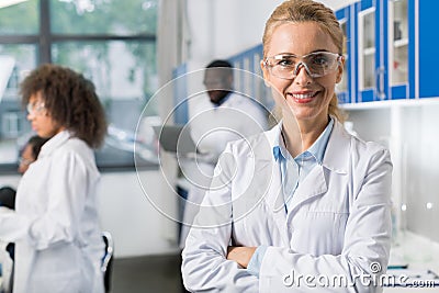 Portrait Of Smiling Woman In White Coat And Protective Eyeglasses In Modern Laboratory, Female Scientist Over Busy Stock Photo