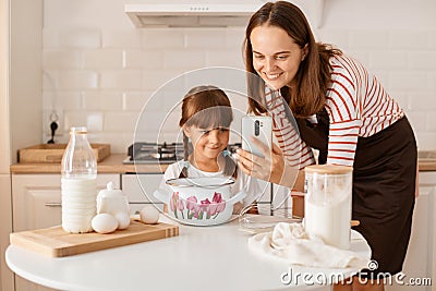 Portrait of smiling young adult woman cooking with her daughter in the kitchen, browsing internet for finding recipe of the pie or Stock Photo