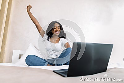 Portrait of smiling young African woman using laptop in bed at home, having video call with her friend, realxing with Stock Photo