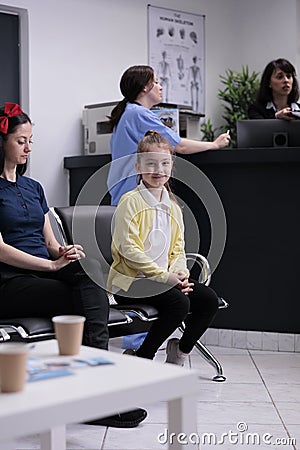 Portrait of smiling young girl with her mother sitting in hospital lobby waiting to see pediatric doctor. Parent with Stock Photo