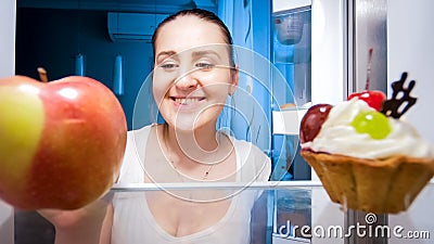 Closeup portrait of smiling young woman doubting between eating apple or cake at night Stock Photo