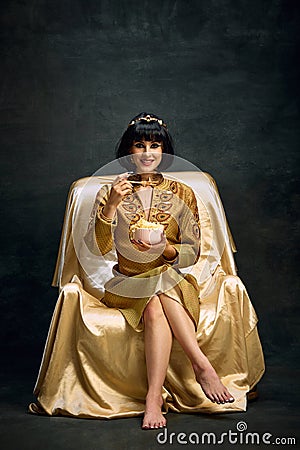 Portrait of smiling young woman in image of egyptian queen, Cleopatra eating instant noodles against dark vintage Stock Photo