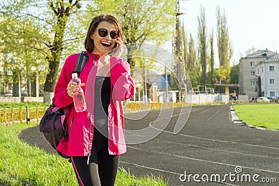 Portrait sporty mature woman at the stadium, in sports clothes for training, with bottle of water, talking on the phone Stock Photo