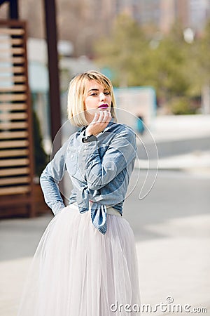 A portrait of a standing dreamy blonde girl with bright pink lips hoding her hand near chin wearing denim shirt, grey Stock Photo