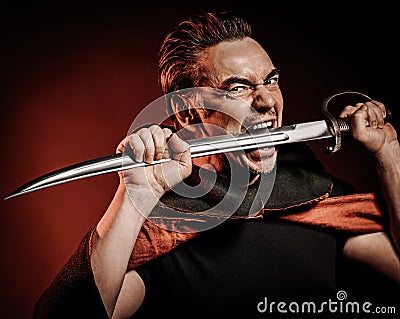 Portrait of strong courage fervor man warrior in cape holding sabre, sword in his mouth, ready to fight over red Stock Photo