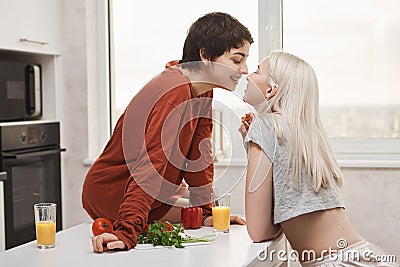 Portrait of stylish and attractive lesbian in red pullover sitting at kitchen table while kissing girlfriend and smiling Stock Photo