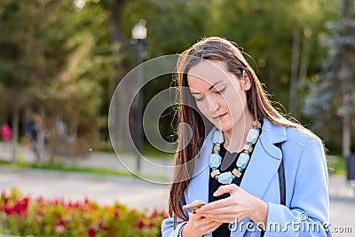 Portrait of stylish girl in a blue coat and beads, reading emails on a smartphone in a Park backlit. Stock Photo