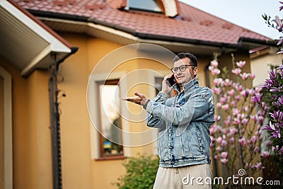 Portrait of stylish man in glasses and jeans jacket, with mobile phone posed against his house and show hand gesture left side Stock Photo