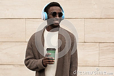 Portrait stylish smiling african man in wireless headphones listening to music with coffee cup wearing brown knitted cardigan Stock Photo