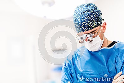 Portrait of surgeon, cosmetic plastician looking down with surgical lamps in background. Doctor with loupes Stock Photo