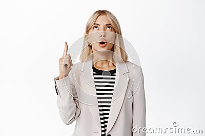 Portrait of surprised blond woman in suit pointing, looking up with fascinated, impressed face expression, checking out Stock Photo