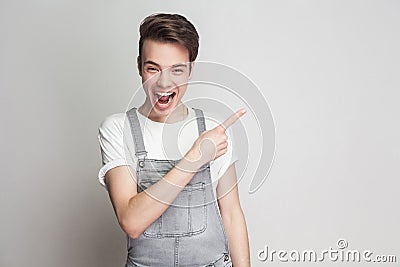 Portrait of surprised young brunette man in casual style with t-shirt and denim overalls standing and looking at camera and point Stock Photo