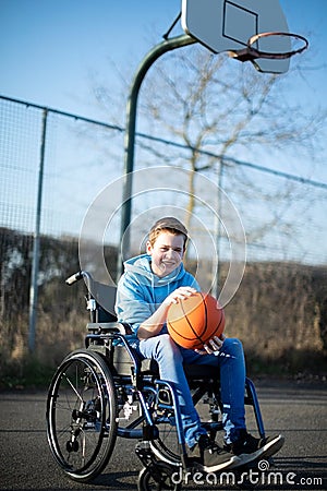 Portrait Of Teenage Boy In Wheelchair Playing Basketball On Outdoor Court Stock Photo