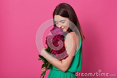 Portrait of tender beautiful young lady wearing in casual green dress, holding dark pink peonies, feeling smell of flowers, having Stock Photo